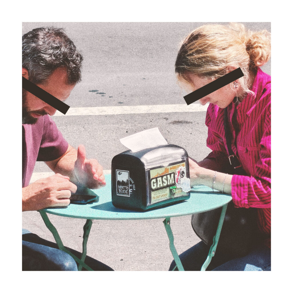 A couple sit at an outdoor table looking at their phones. A napkin dispenser in front of them has a sticker with the name GASM.
