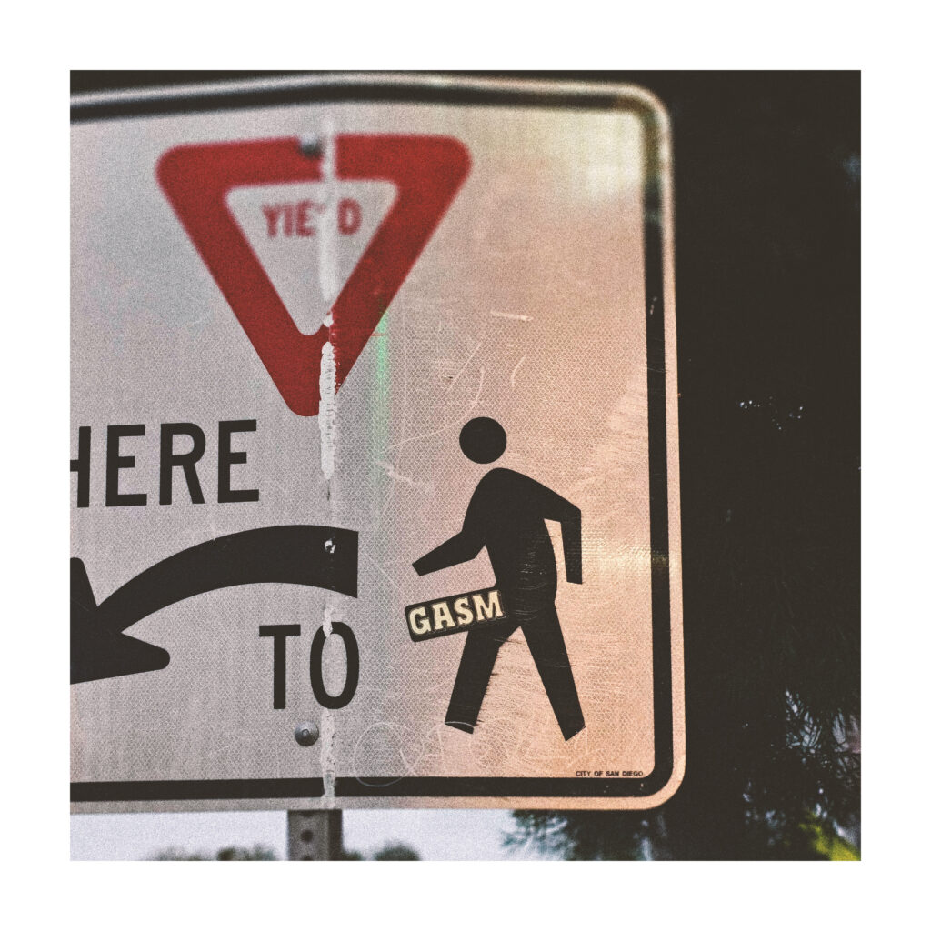 A yield to pedestrians sign that has a sticker with the name GASM placed on the person icon of the sign, emulating a penis.